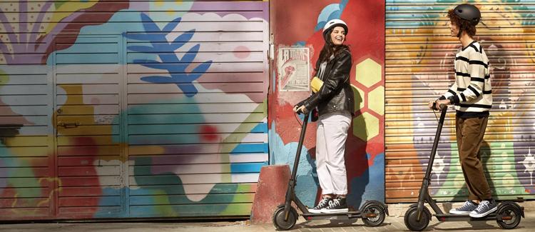 En jente og en gutt med hjelm på el sparkesykkel foran en fargerik vegg.
